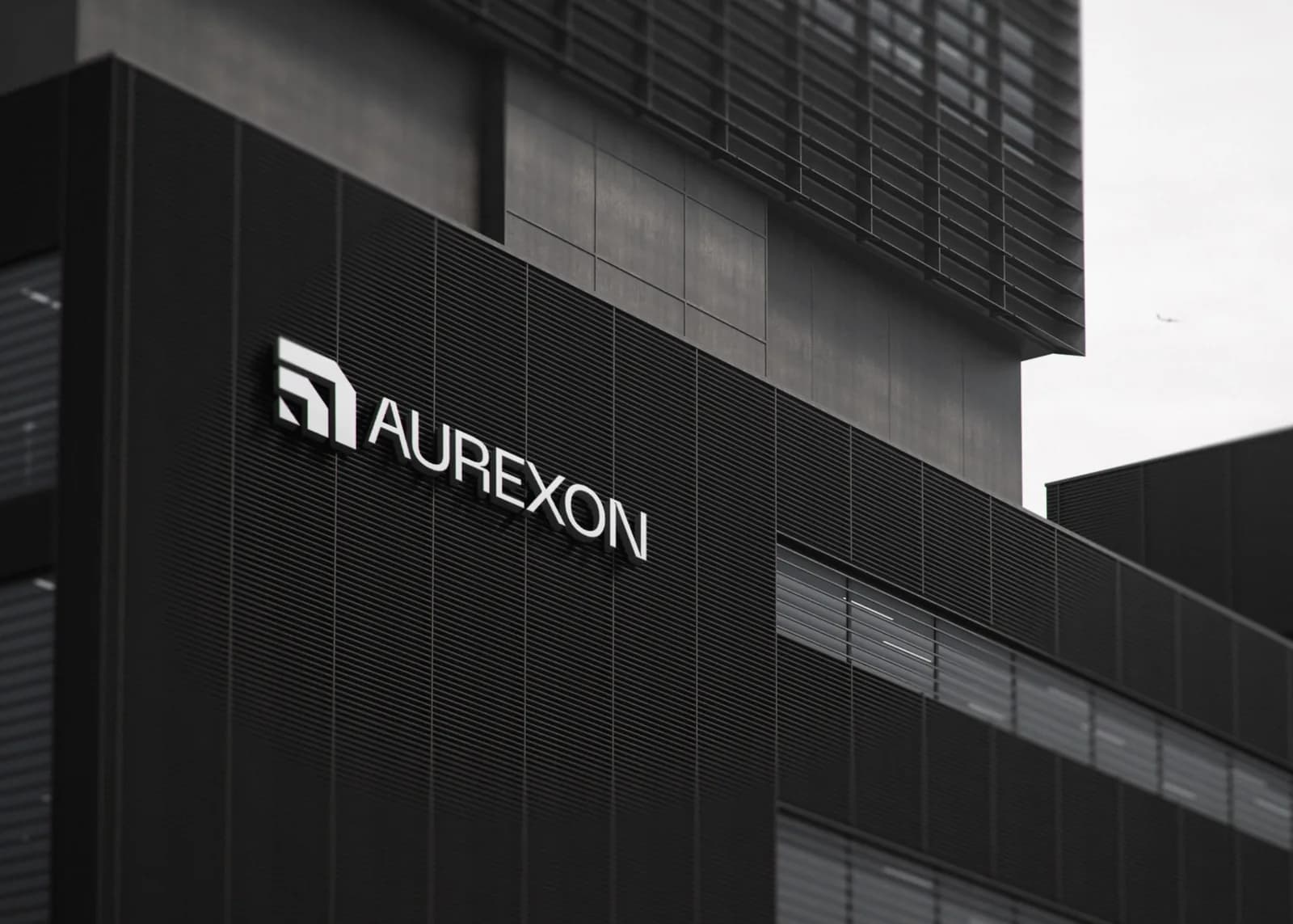 The dimensional Aurexon symbol and wordmark in white, mounted on the dark ribbed facade of a modern office building.