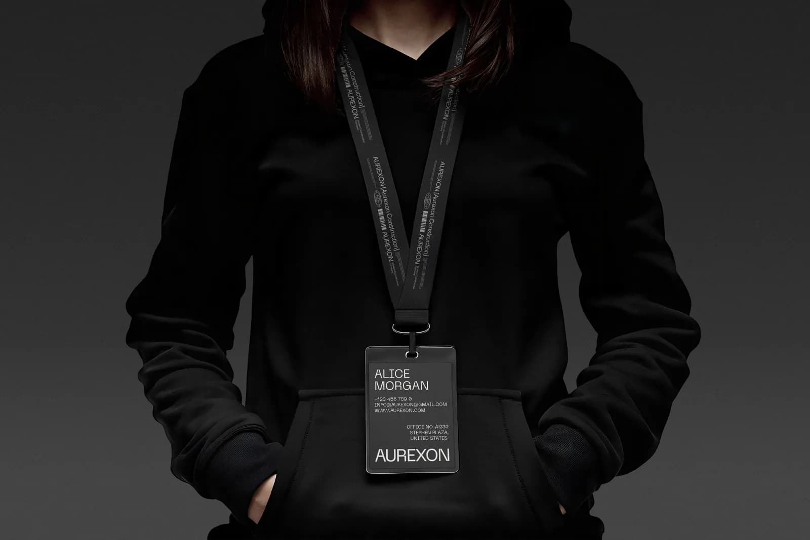 A person in a black hoodie wearing a black lanyard printed with AUREXON [Aurexon Construction], holding a dark ID badge with the name Alice Morgan and the AUREXON wordmark.